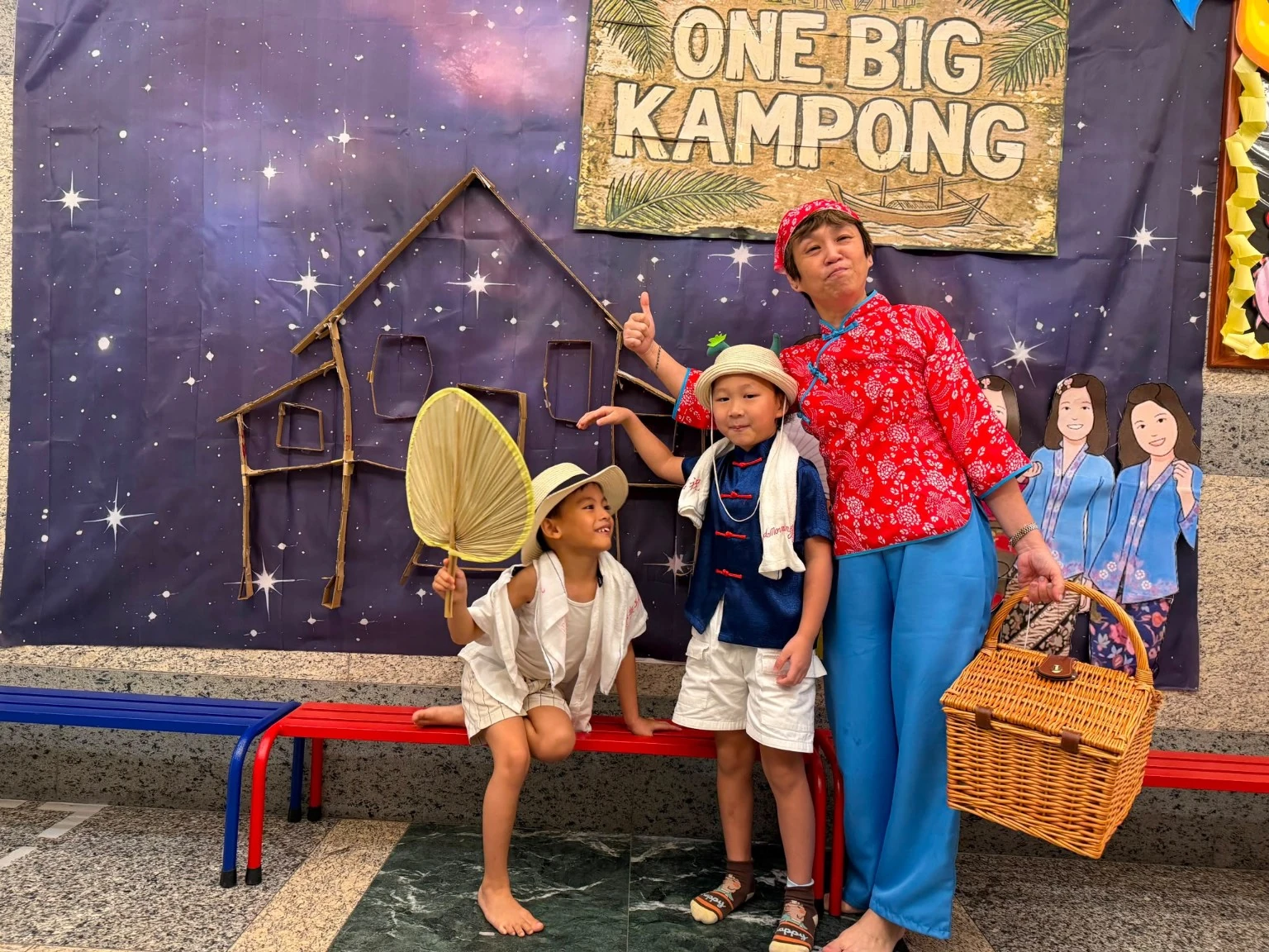 An educator and two children in themed clothing beside a One Big Kampong display.