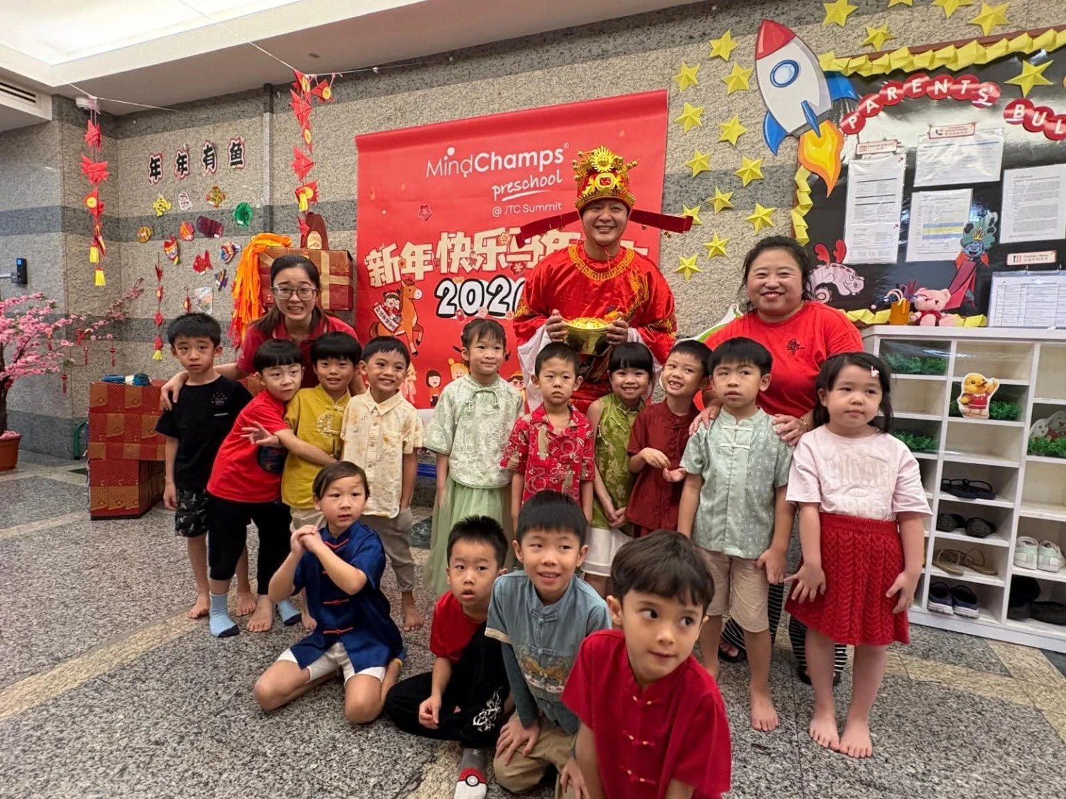 Children and educators in festive clothing gathered beside a MindChamps JTC Summit celebration display.